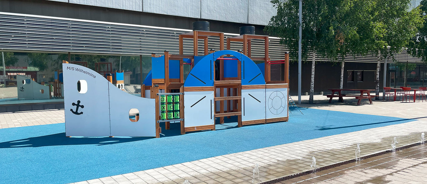 Lekbåt på blått underlag med hus i bakgrunden och vattenfontäner i förgrunden.