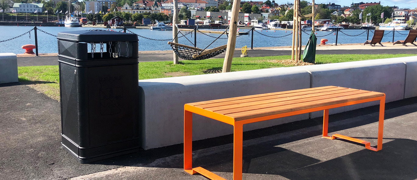 Orange parkbänk och svart papperskorg på en strandväg vid havet.