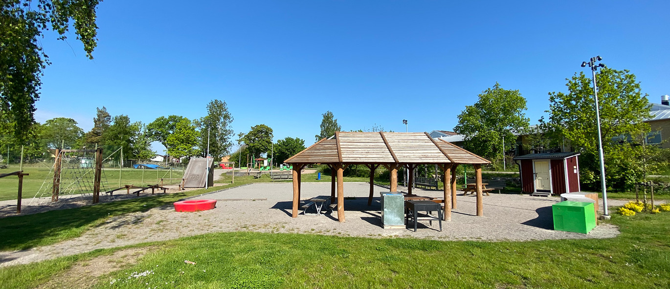 Solig dag i parken med grillhus, hinderbana och grönt gräs.