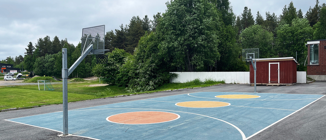 Färgrik basketplan på asfalt, med basketstativ och träd och grönska i bakgrunden.