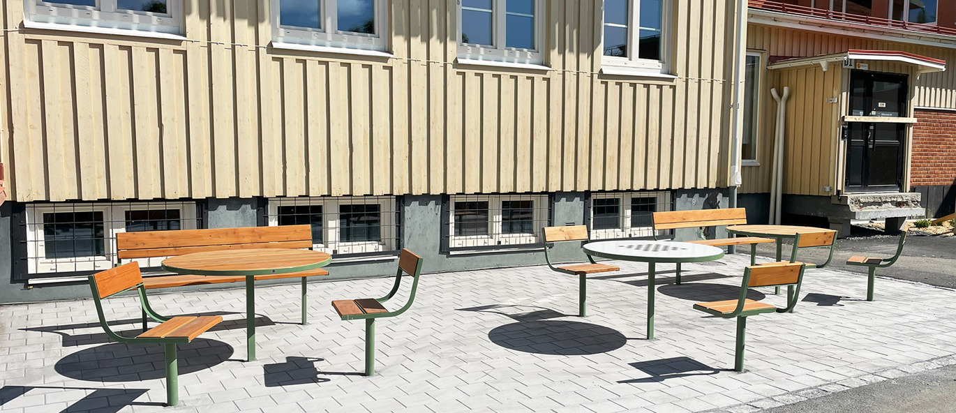 Utemöbler i trä och grönt stål på skolgård med marksten och gul skolbyggnad i bakgrunden.