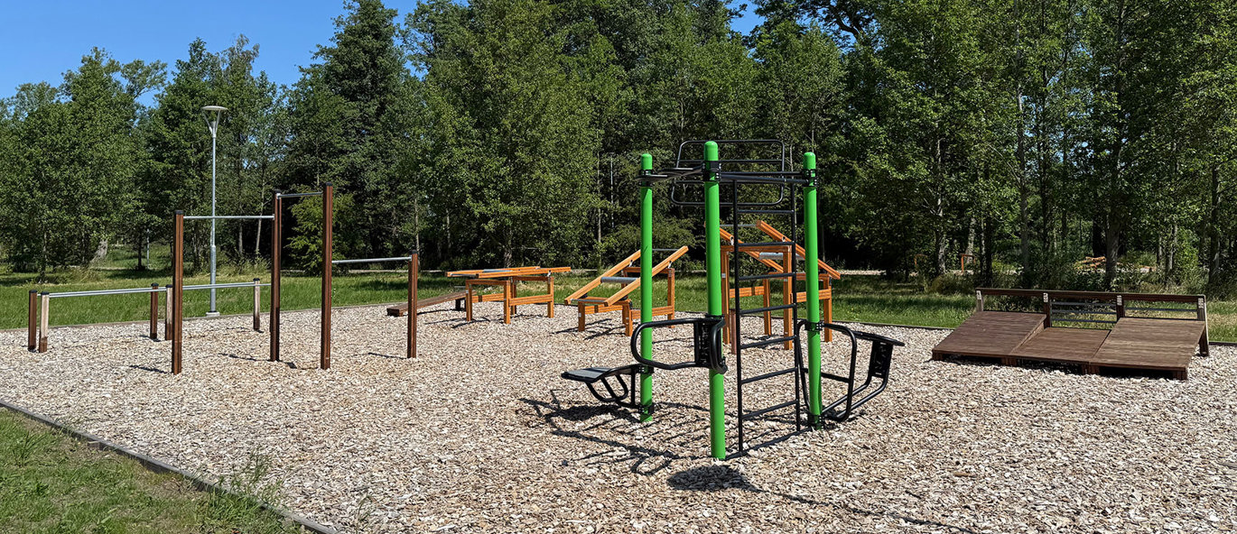 Grön träningsställning samt flera träningsstationer av trä på flisunderlag, med grönt gräs och träd i bakgrunden