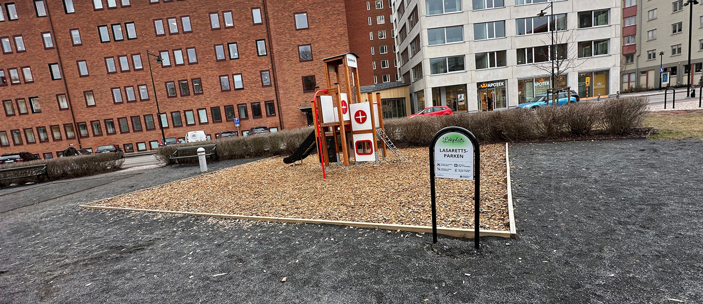 Lekställning och informationsskylt om lekparken framför stora byggnader.