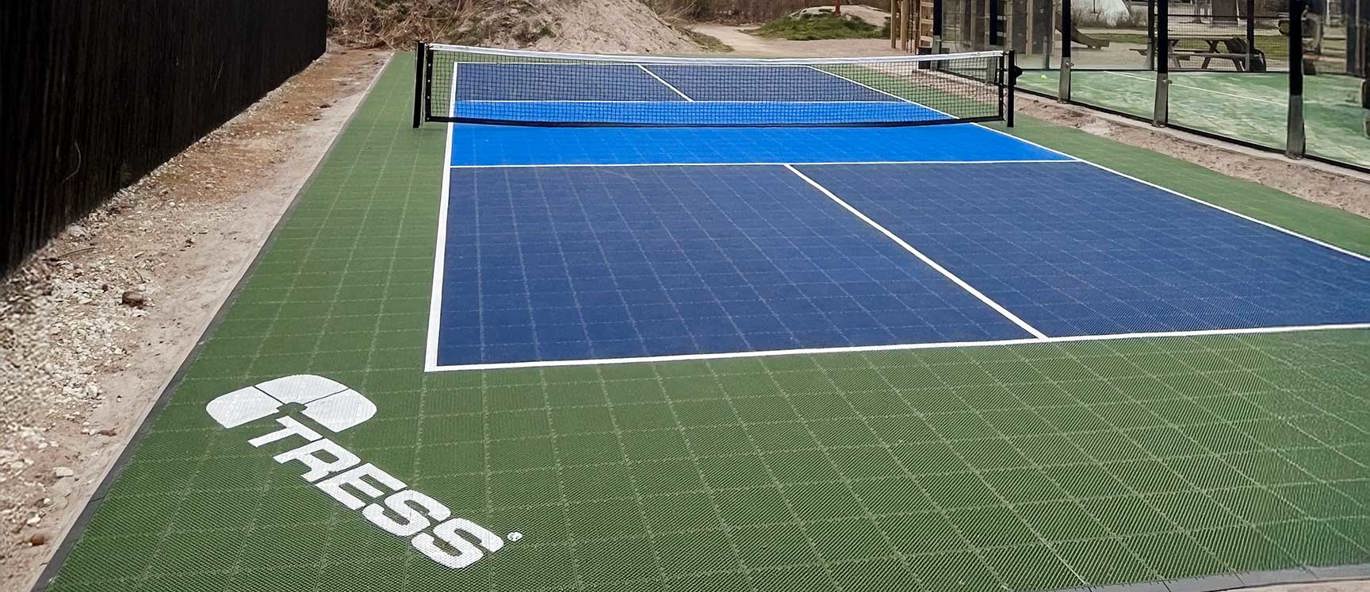 Blå pickleballbana med blått nät på grönt underlag med Tress logo i vitt och en padelbana i bakgrunden.
