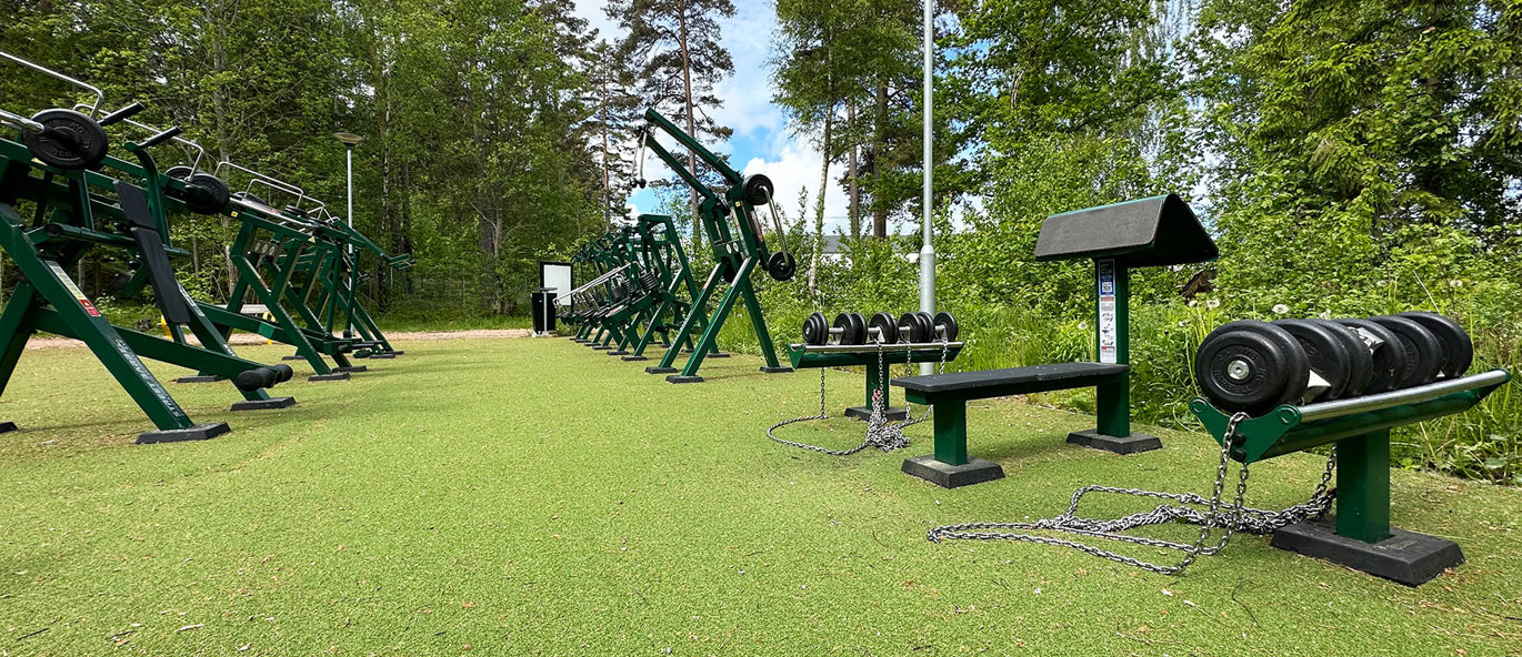 Grönt konstgräs, mossgröna träningsmaskiner och skog i bakgrunden.