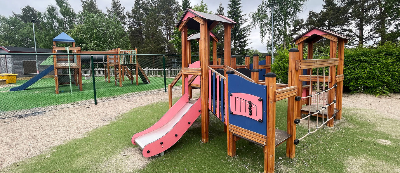 Färgglad klätterställning av trä som står på ett grönt konstgräs och en annan klätterställning i bakgrunden.