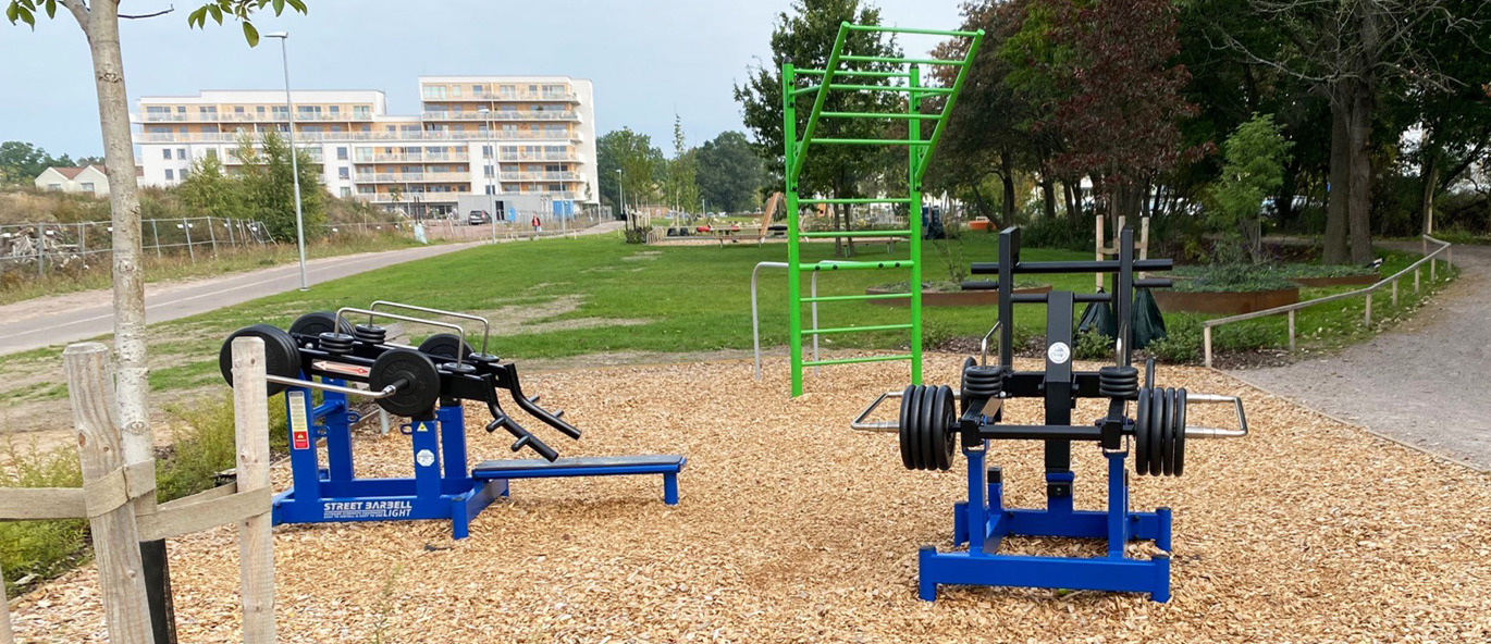 Två blåa träningsmaskiner och en grön träningsställning i en park med flis som underlag.