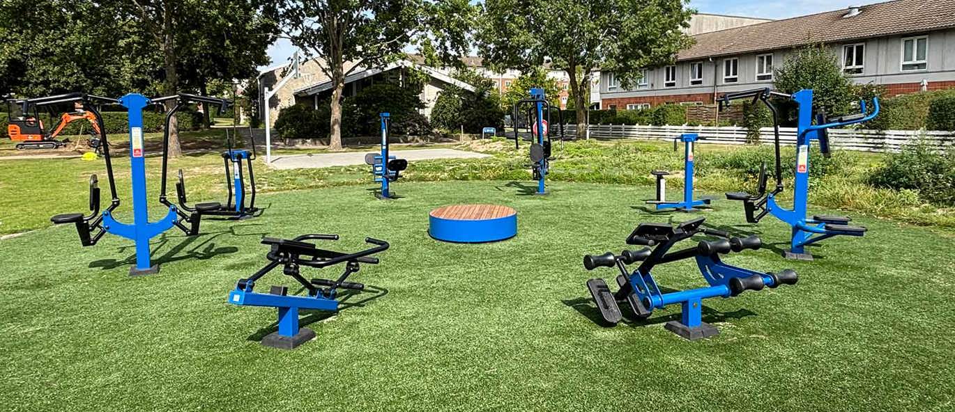 Grön gräsmatta med blå träningsstationer i en cirkel med bostadshus i bakgrunden.