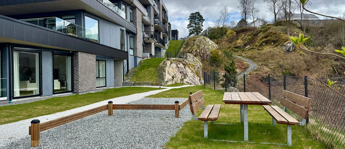 Bänkbord och balansbom av trä, grönt gräs, grusplan och bostadshus med berg i bakgrunden.